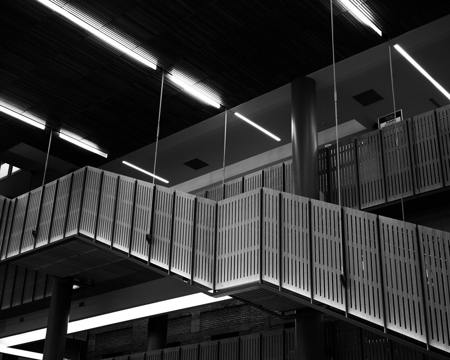 Industrial ceiling with fluorescent lights and suspended walkway featuring metal railings in monochrome.