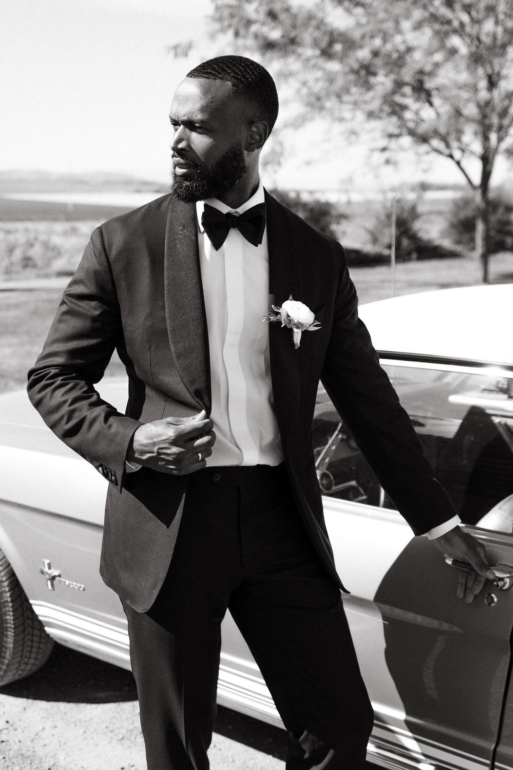 Elegant groom in black tuxedo with bow tie and boutonniere leaning against classic car in black and white photo.