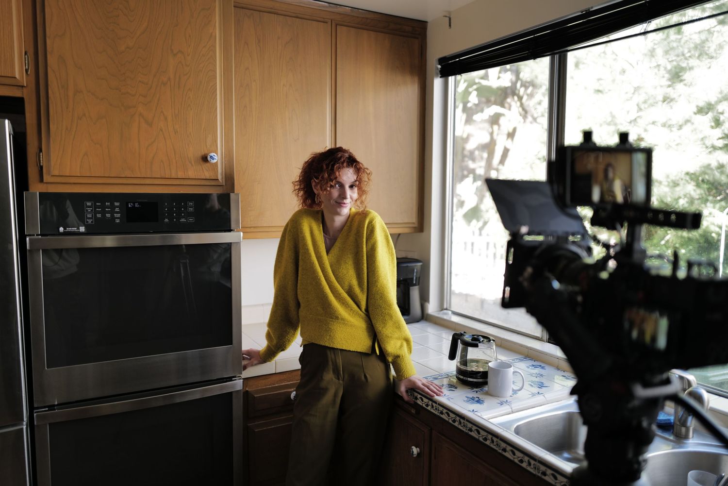 Woman in yellow sweater standing in modern kitchen being filmed by a camera near a window.
