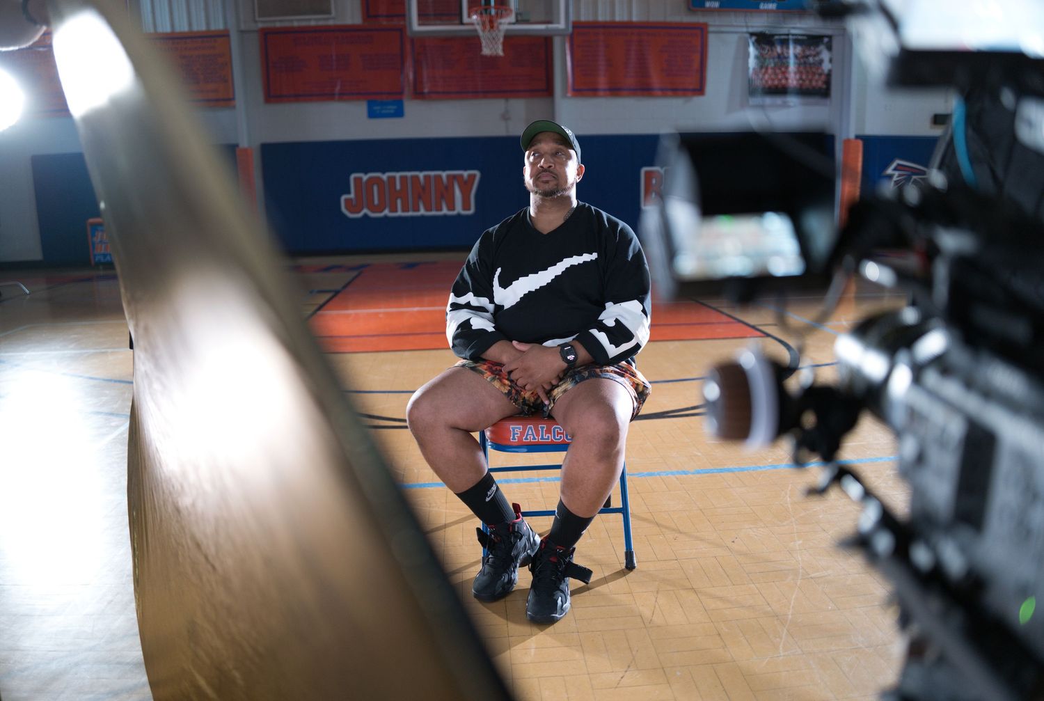 Athlete sits on chair in gym during Nike video shoot, with cameras and basketball court visible in background.