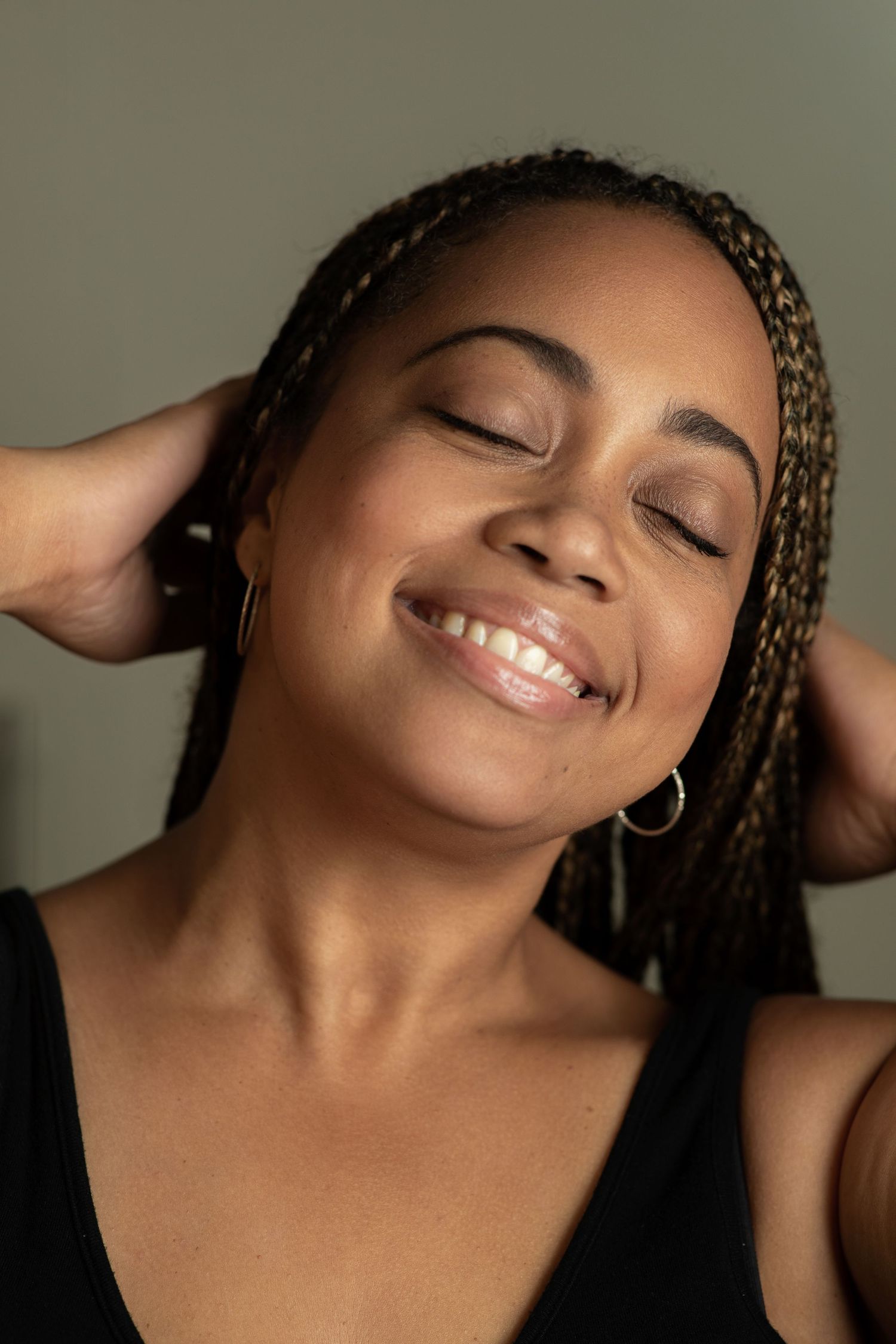 A woman with braids smiling with closed eyes and hands behind head.