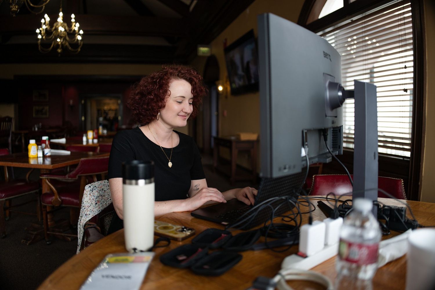 Woman with curly red hair working on desktop computer at wooden desk in cozy restaurant setting.