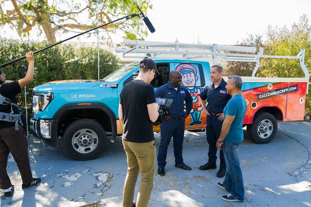 Film crew shooting a commercial with actors near a colorful service truck outdoors.