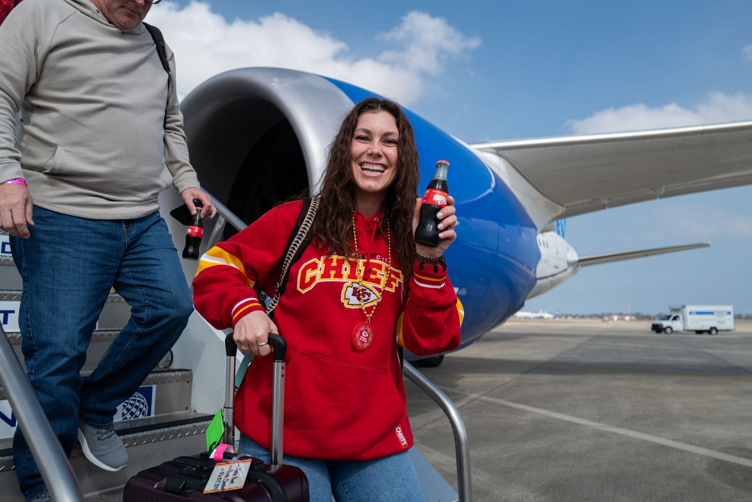 A fan in a red Chiefs sweatshirt poses with a drink next to an airplane engine on the tarmac.