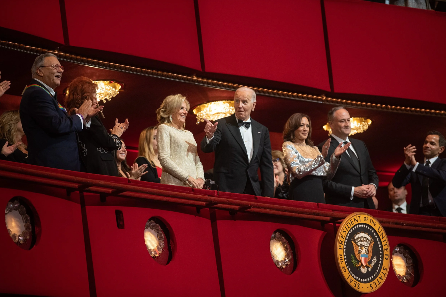 Audience members in formal attire stand and applaud from a theater balcony with red decor and presidential seal visible.