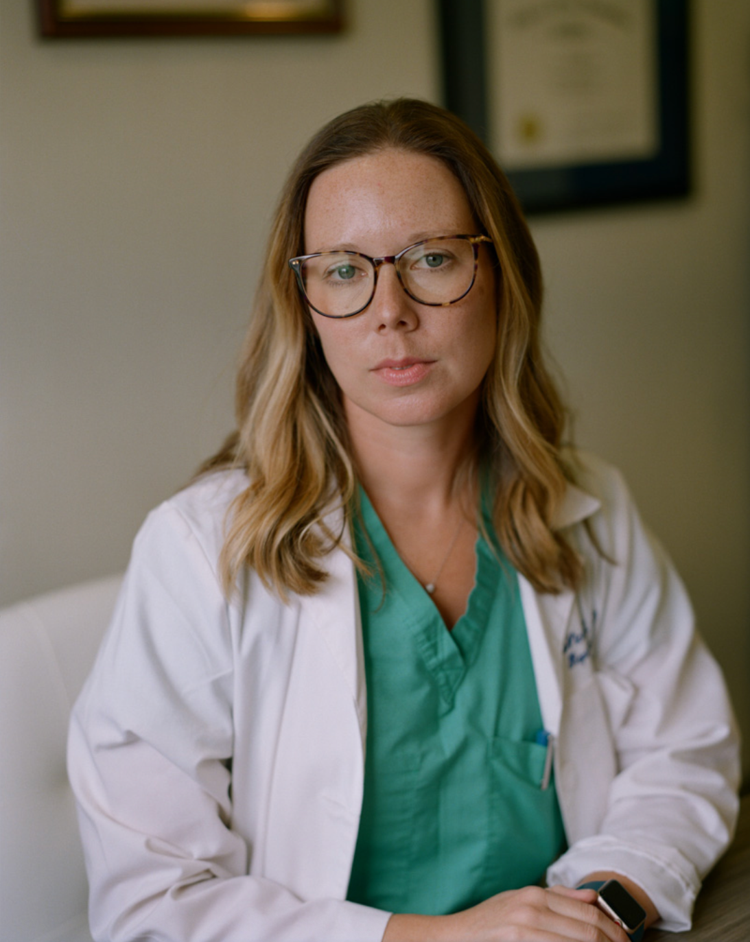 Healthcare professional in white coat and teal scrubs writes notes at desk with framed artwork on wall behind.