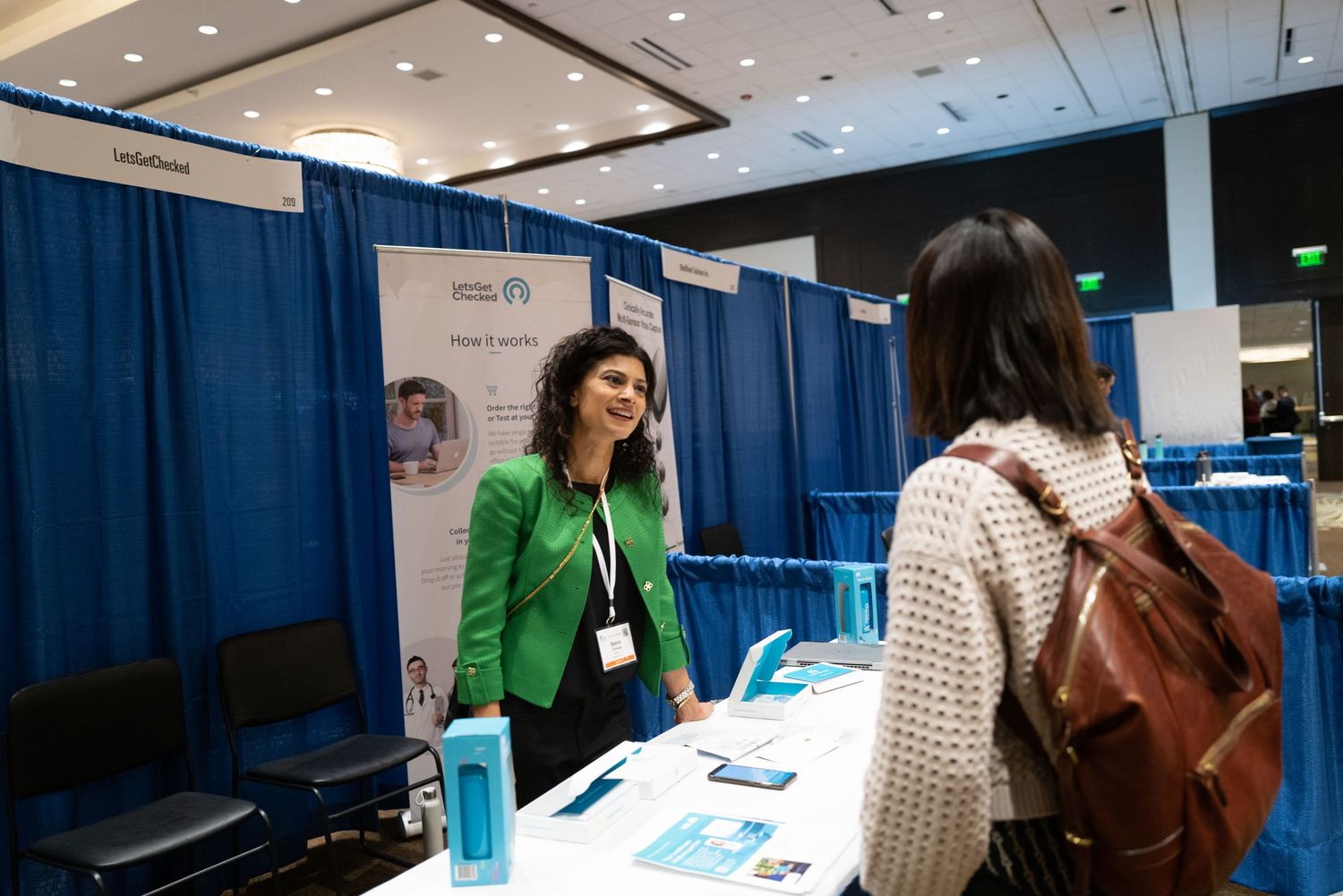 Trade show booth with exhibitor in green jacket speaking to attendee.