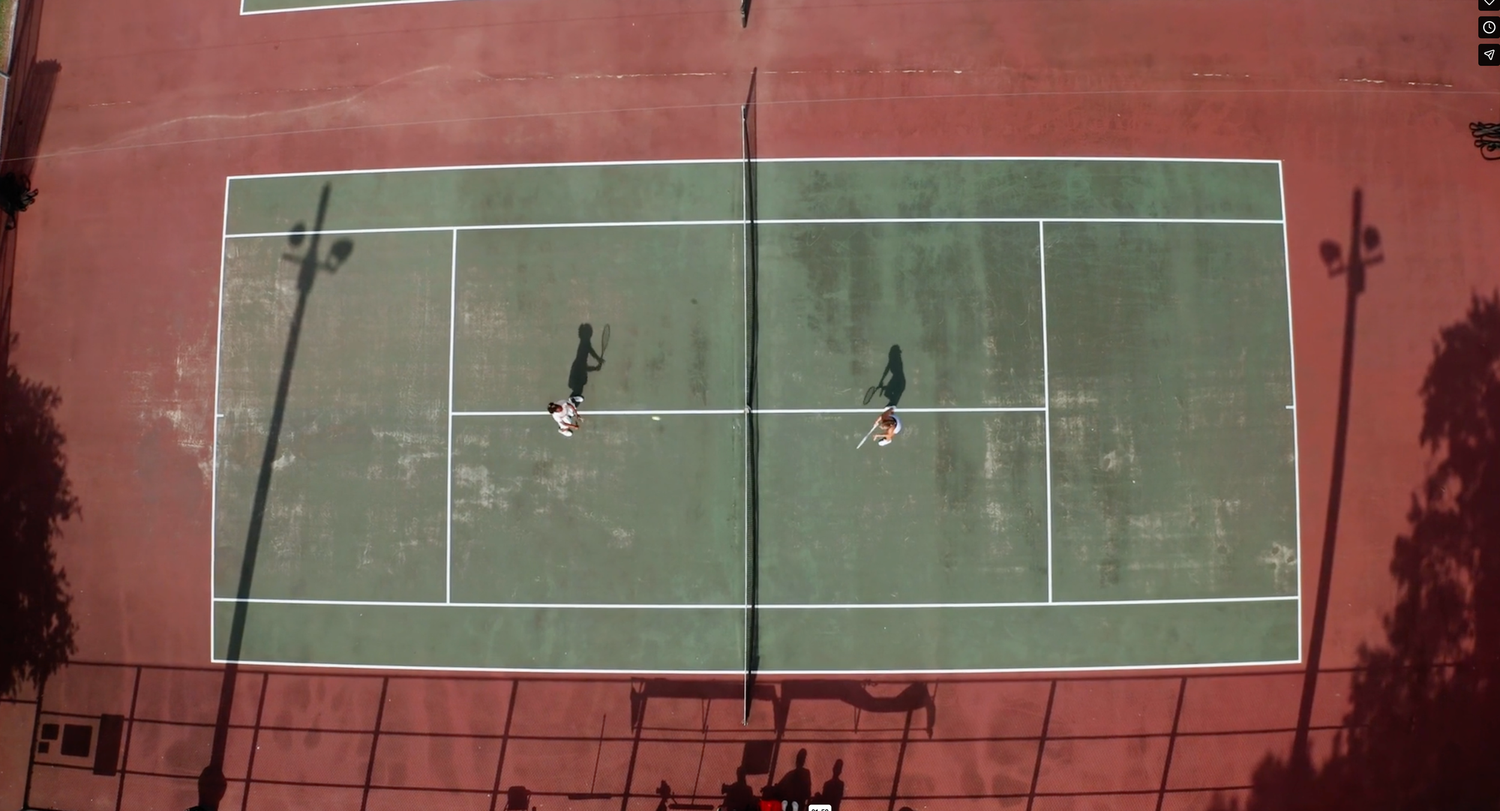 Aerial view of two players on a green and red outdoor tennis court with long shadows cast across the surface.
