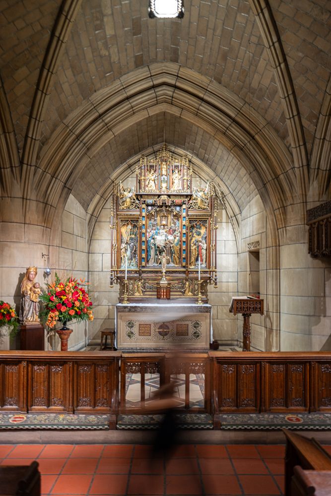 Interior view of a Gothic church altar with ornate golden reredos, stone archway, and red tiles under wooden railing.