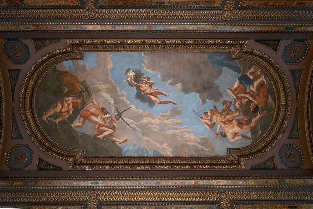 Ornate baroque fresco paintings on a palace ceiling depict mythological scenes with classical figures and cherubs in golden frames.