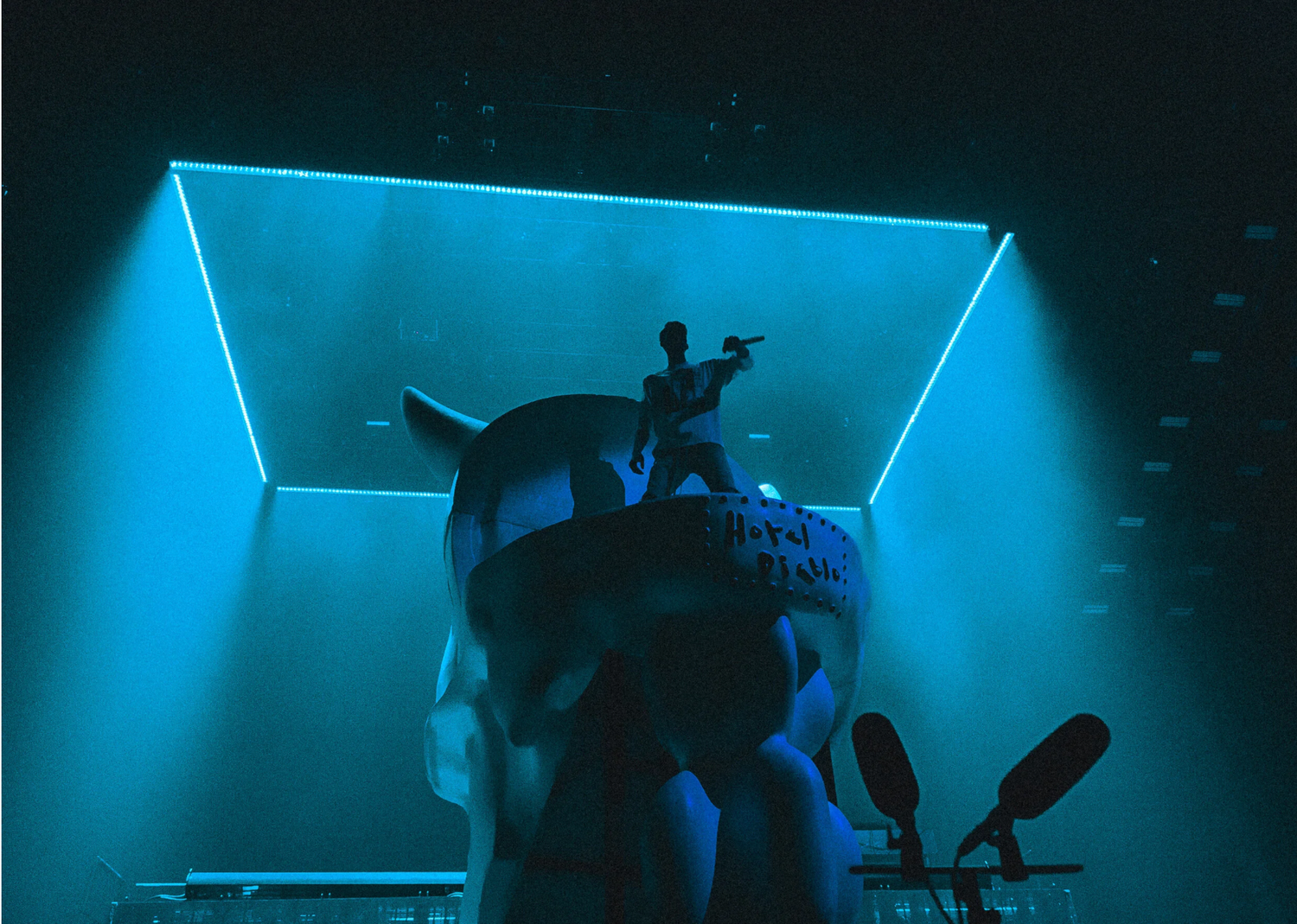 A performer silhouetted on stage with dramatic blue neon lighting and geometric shapes in the background.