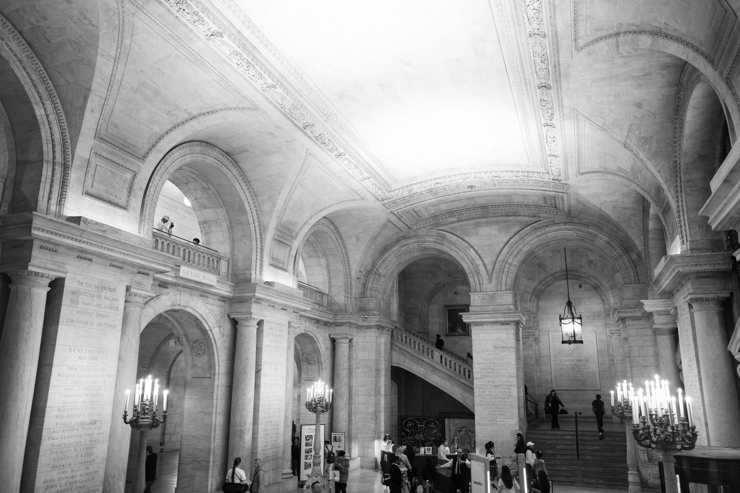 Black and white photo of grand interior hall with arched ceilings, ornate columns, chandeliers, and a sweeping staircase.