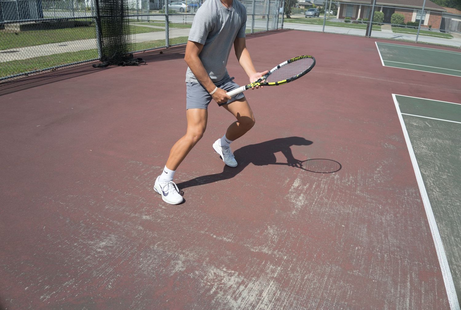 Man playing tennis on an outdoor court, mid-swing with a racket on a red hard court surface.