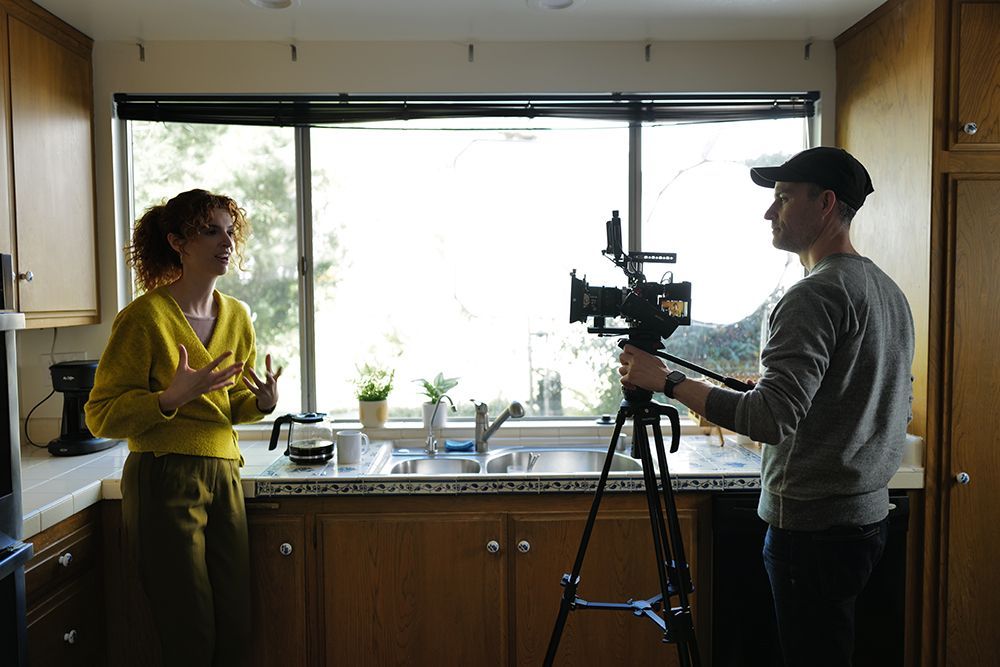 Videographer filming a woman in a yellow top during an interview in a modern kitchen setting.