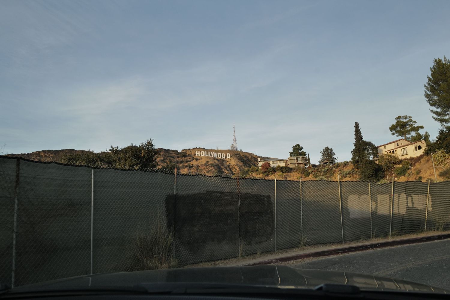 View from a car of the Hollywood Sign on a hilltop behind a chain-link fence in Los Angeles, CA.