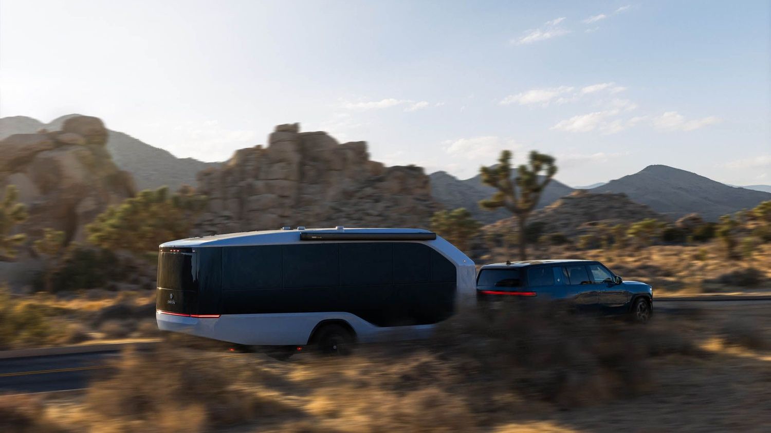 A modern electric trailer being towed by a pickup truck on a desert highway surrounded by rocky terrain and Joshua trees.