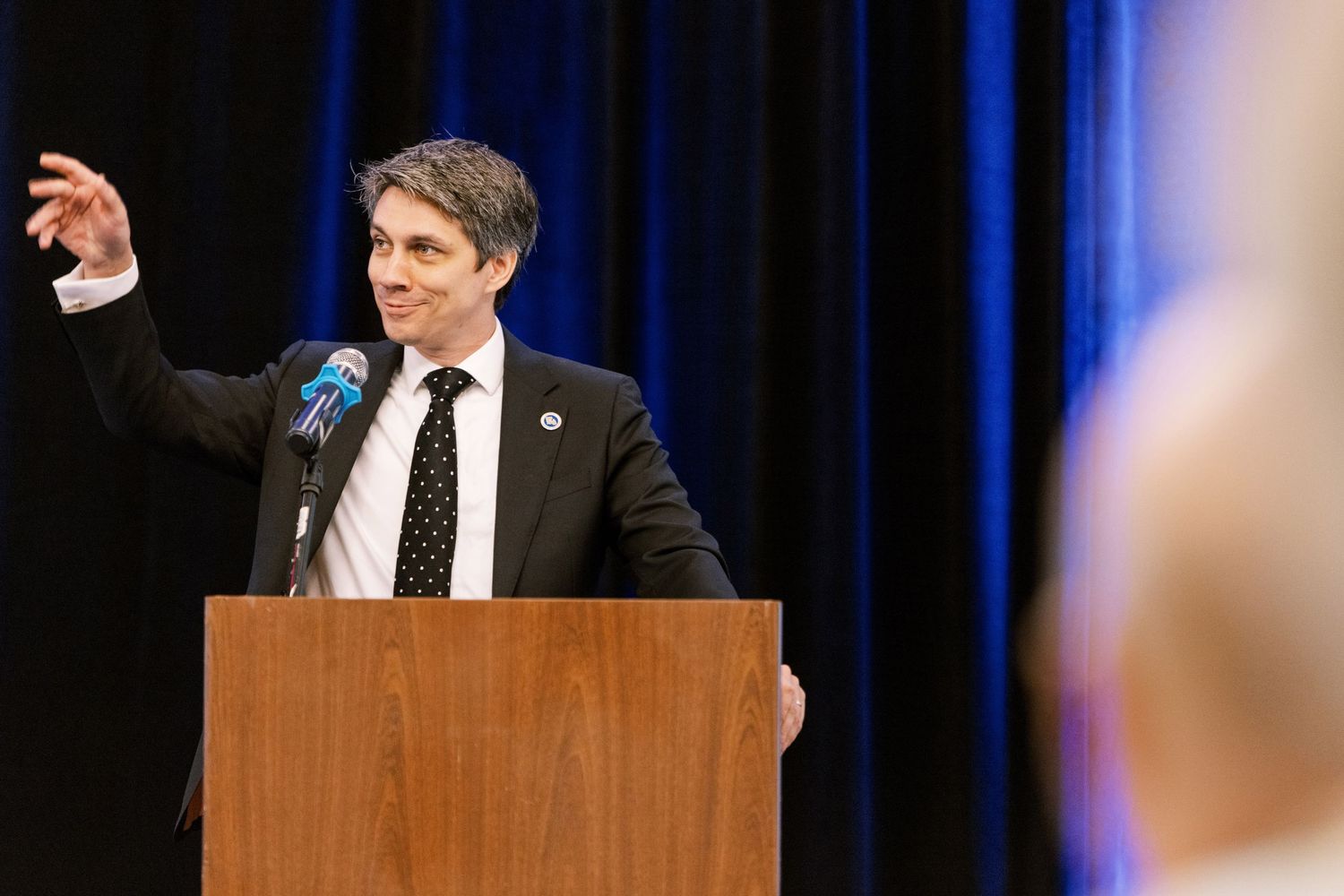 A speaker gestures while delivering remarks at a wooden podium on stage with dark blue curtains.