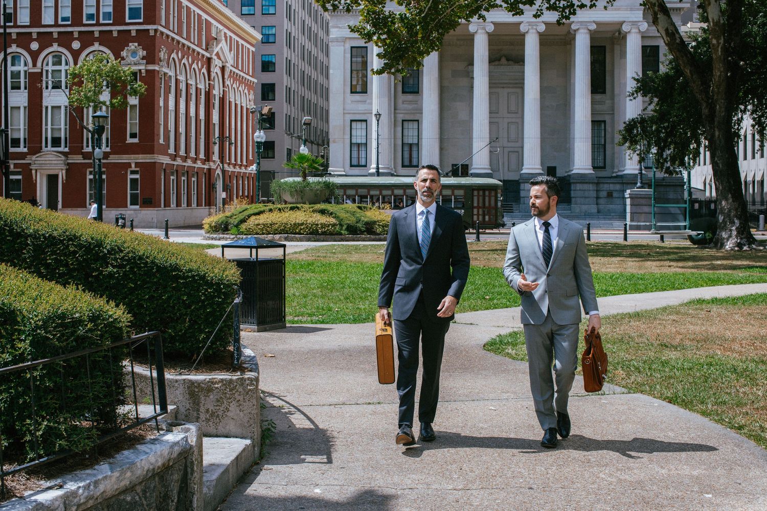 Students walking on a college campus path between brick buildings and green lawns on a sunny day.