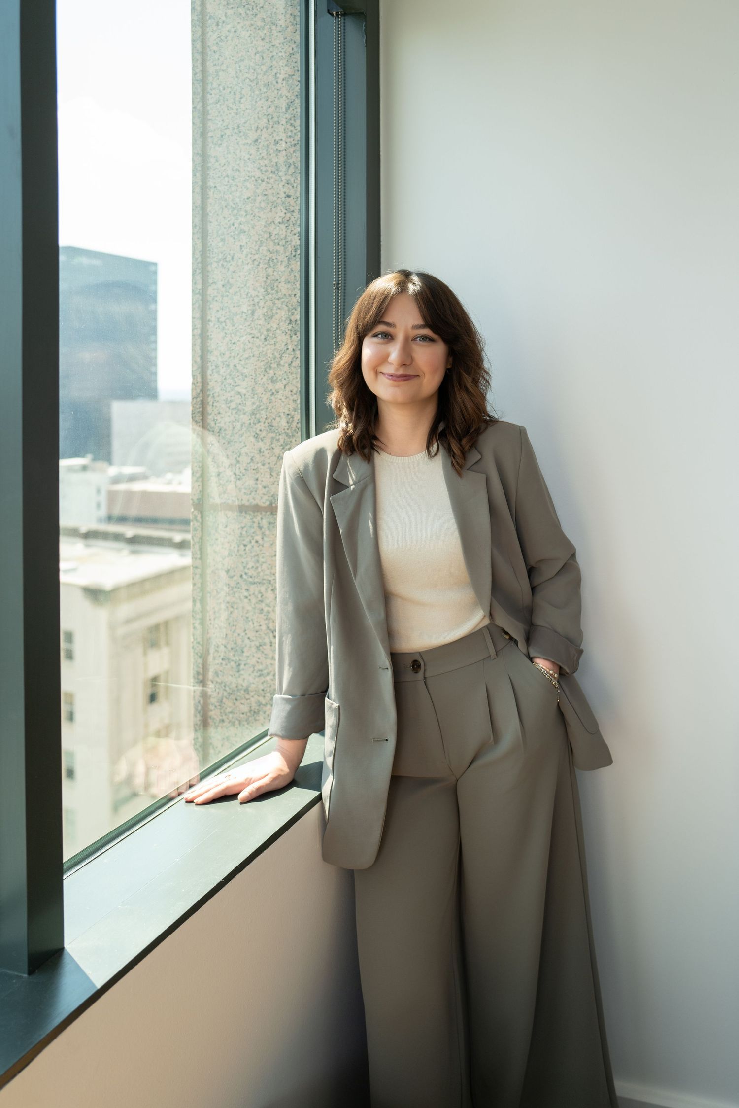 Professional in a gray business suit poses by bright office windows with modern interior and city views.