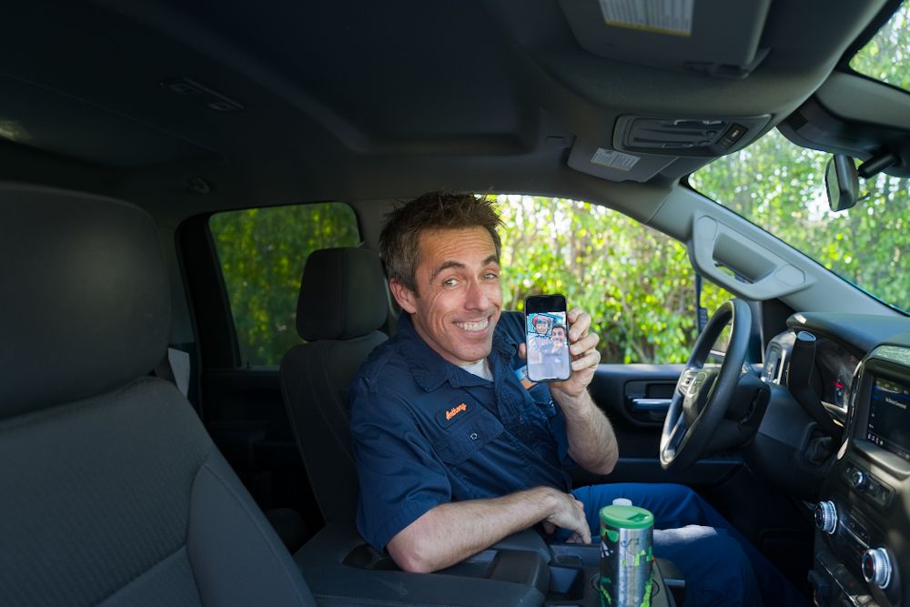 A smiling man in a blue uniform shirt holds up a smartphone while sitting in a pickup truck.