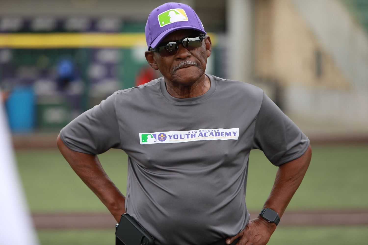 Baseball coach in purple cap and grey shirt on field.