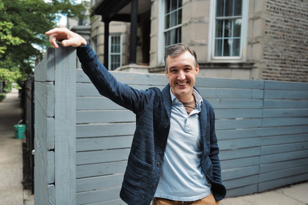 Smiling middle-aged man leaning on a gray wooden fence outdoors on a city street.