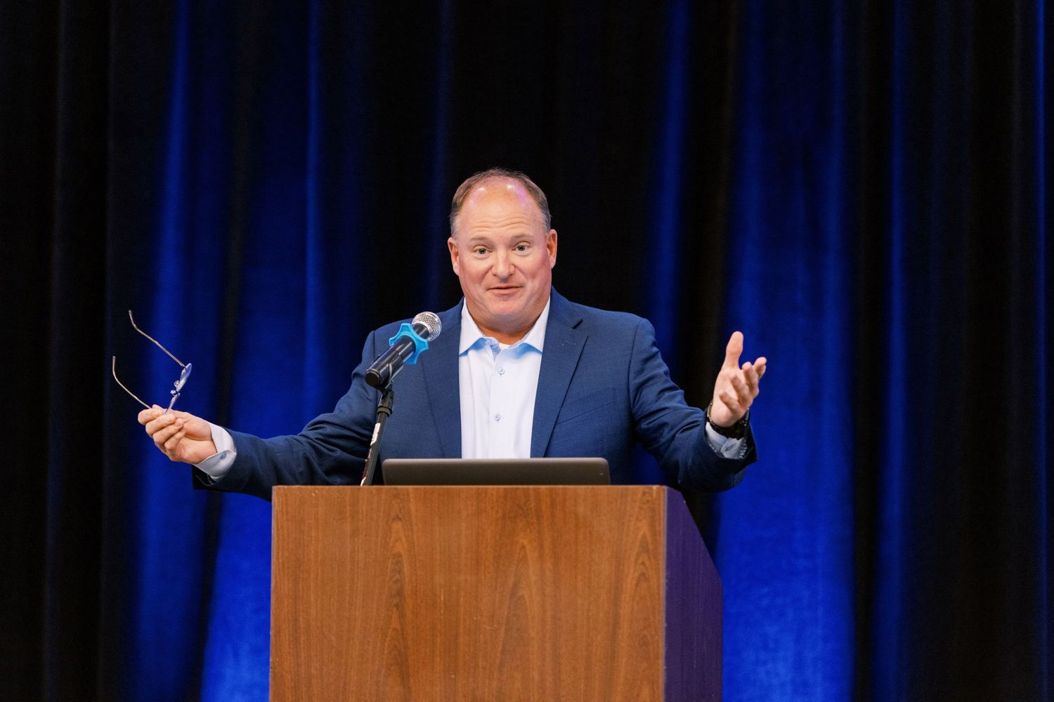 A speaker presents at a wooden podium against blue curtains during a professional conference.