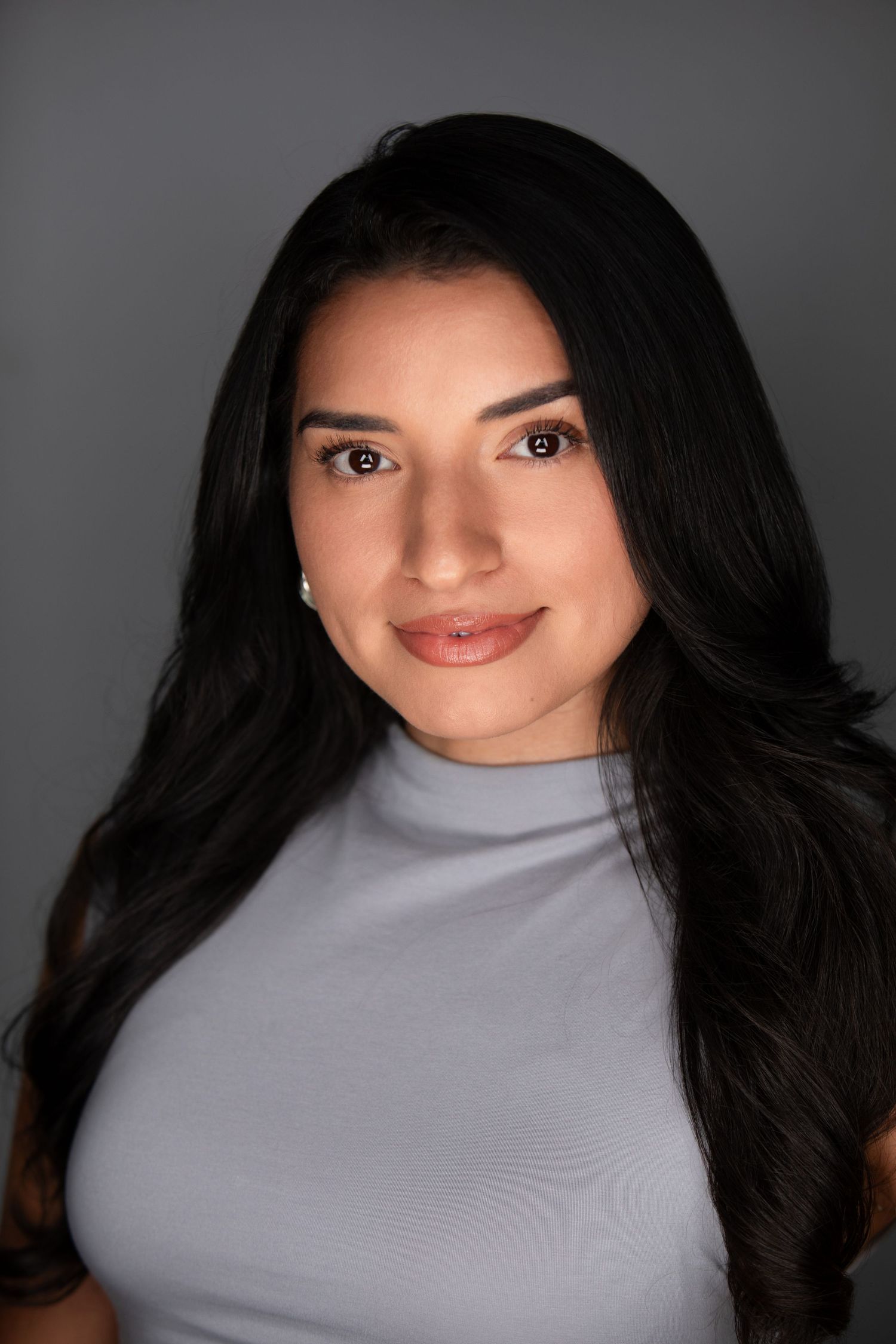 A young woman with long dark hair wearing a gray turtleneck top poses for a professional headshot photo.