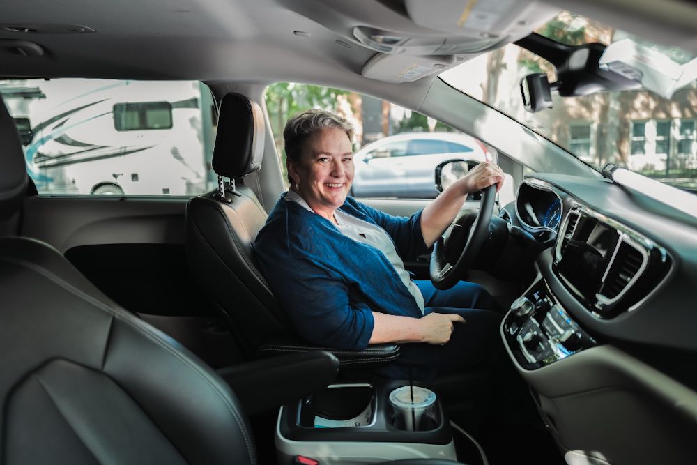 Smiling middle-aged woman in blue top driving a modern car with leather interior, hands on steering wheel.