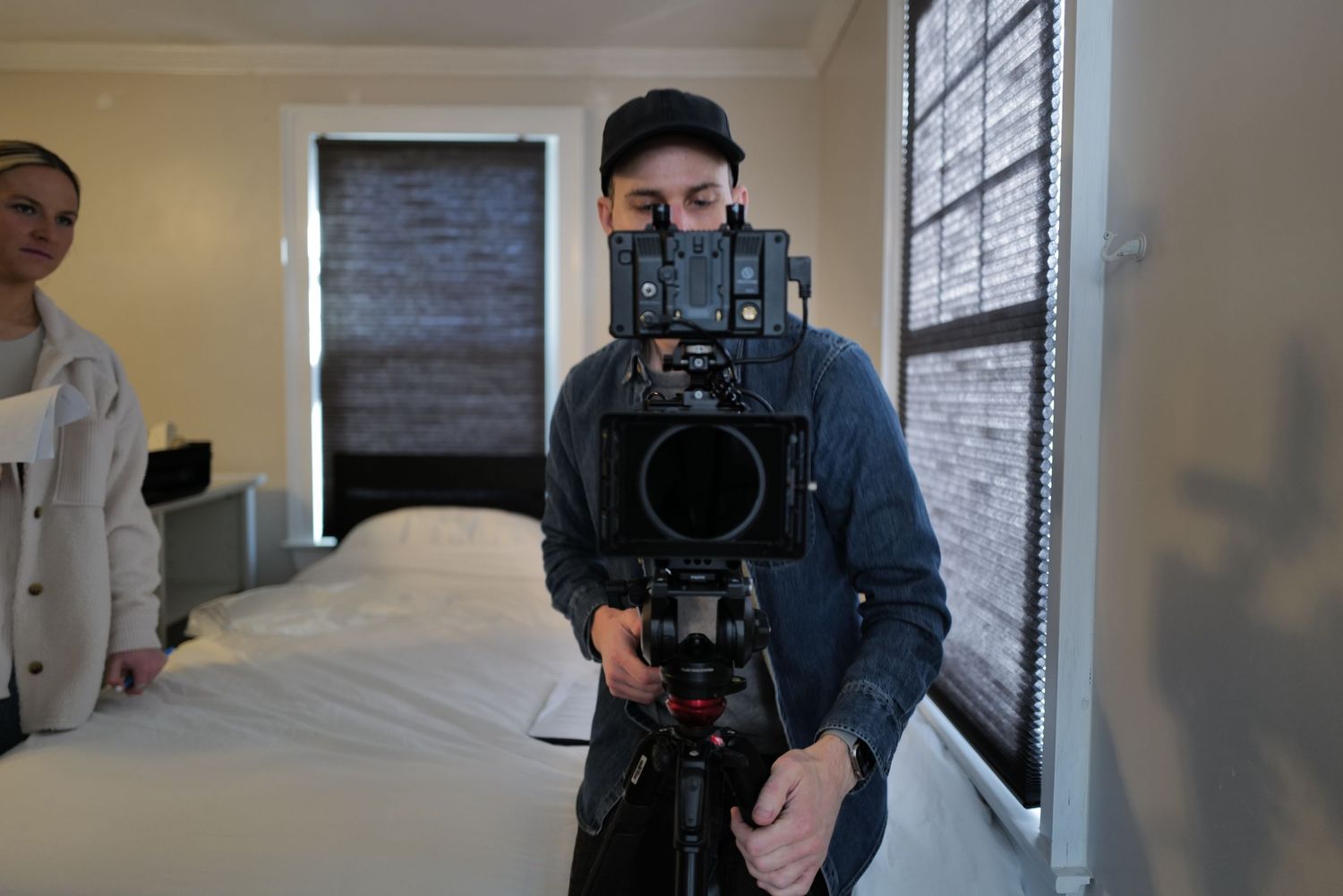 Filmmaker operating a professional cinema camera on a tripod in a bedroom, with a woman standing nearby.
