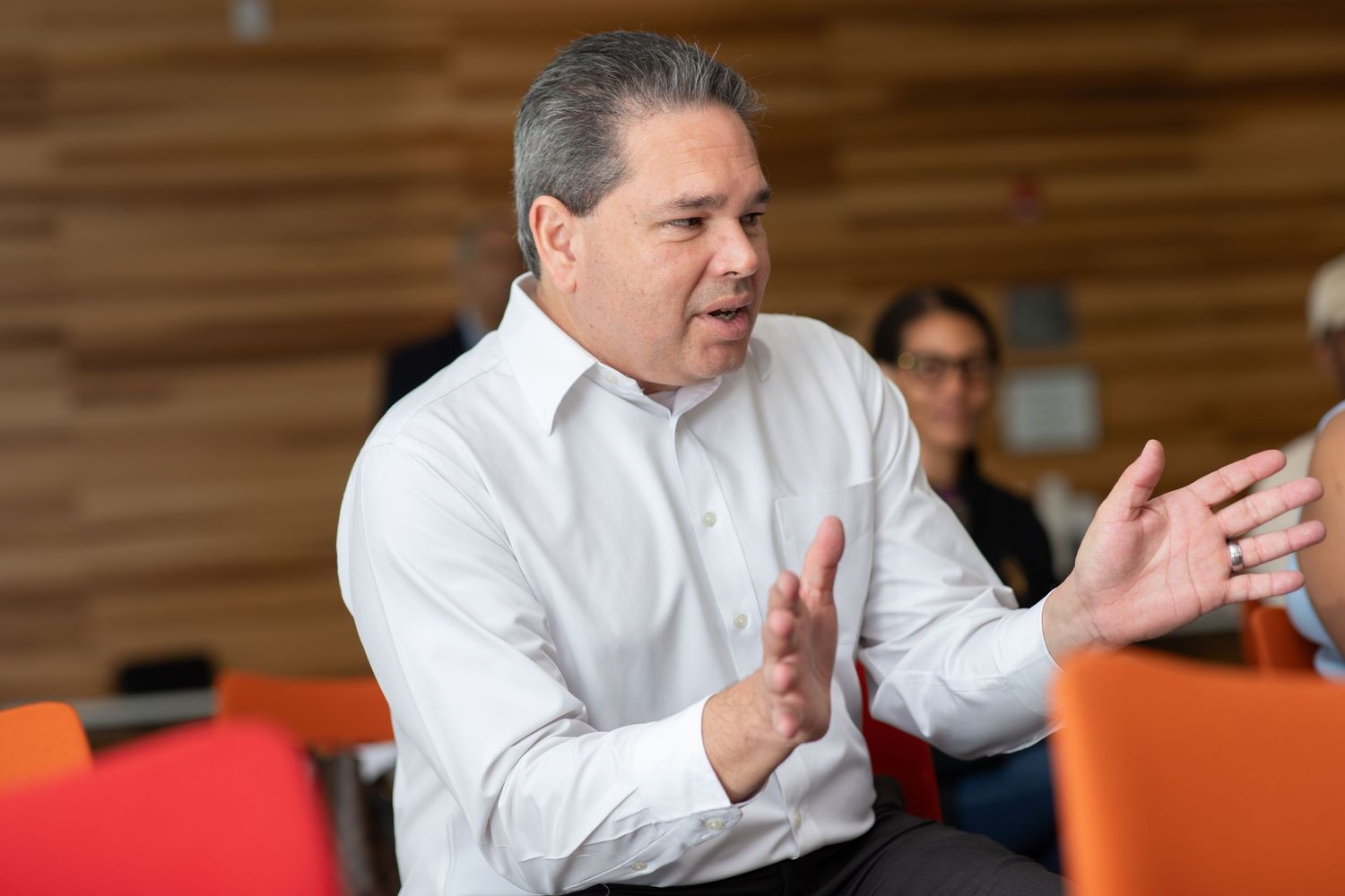 A person in a white dress shirt gestures while speaking in an office setting with orange chairs.