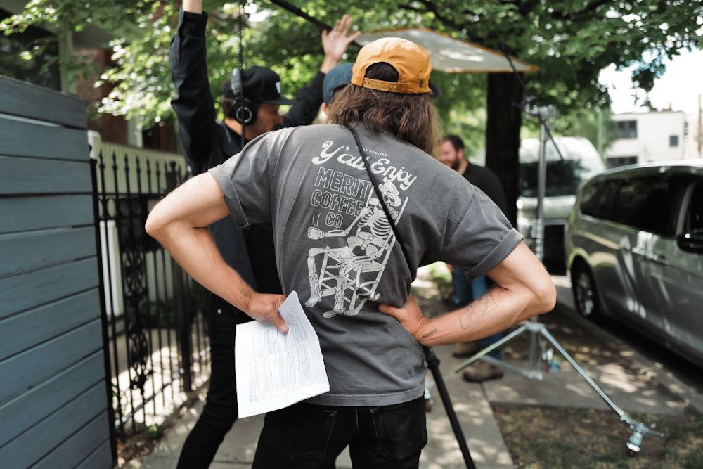 Man in gray graphic t-shirt holds papers outdoors on a film set with crew members and equipment visible.