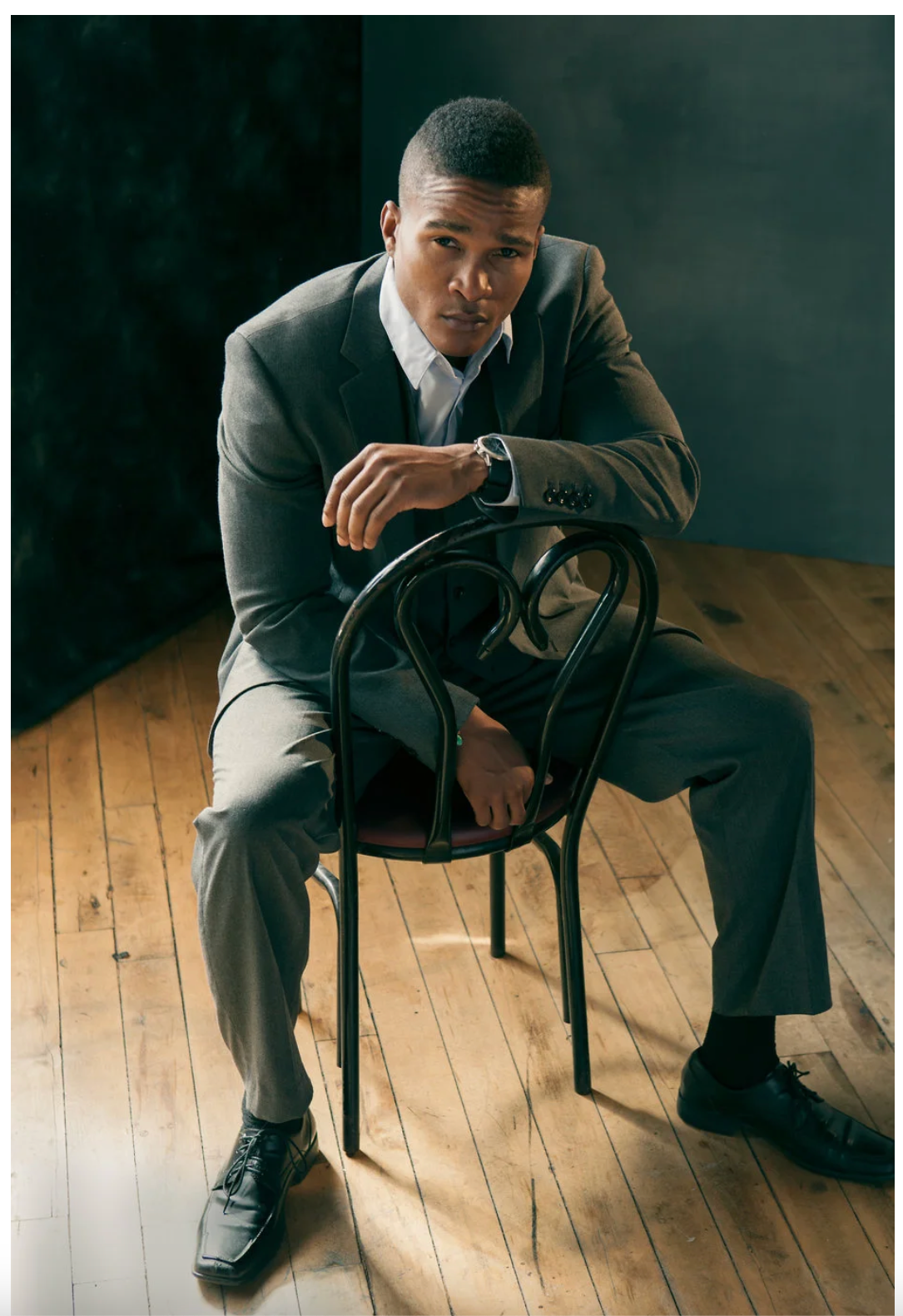 Professional portrait of man in grey suit sitting backwards on wooden chair against dark background.