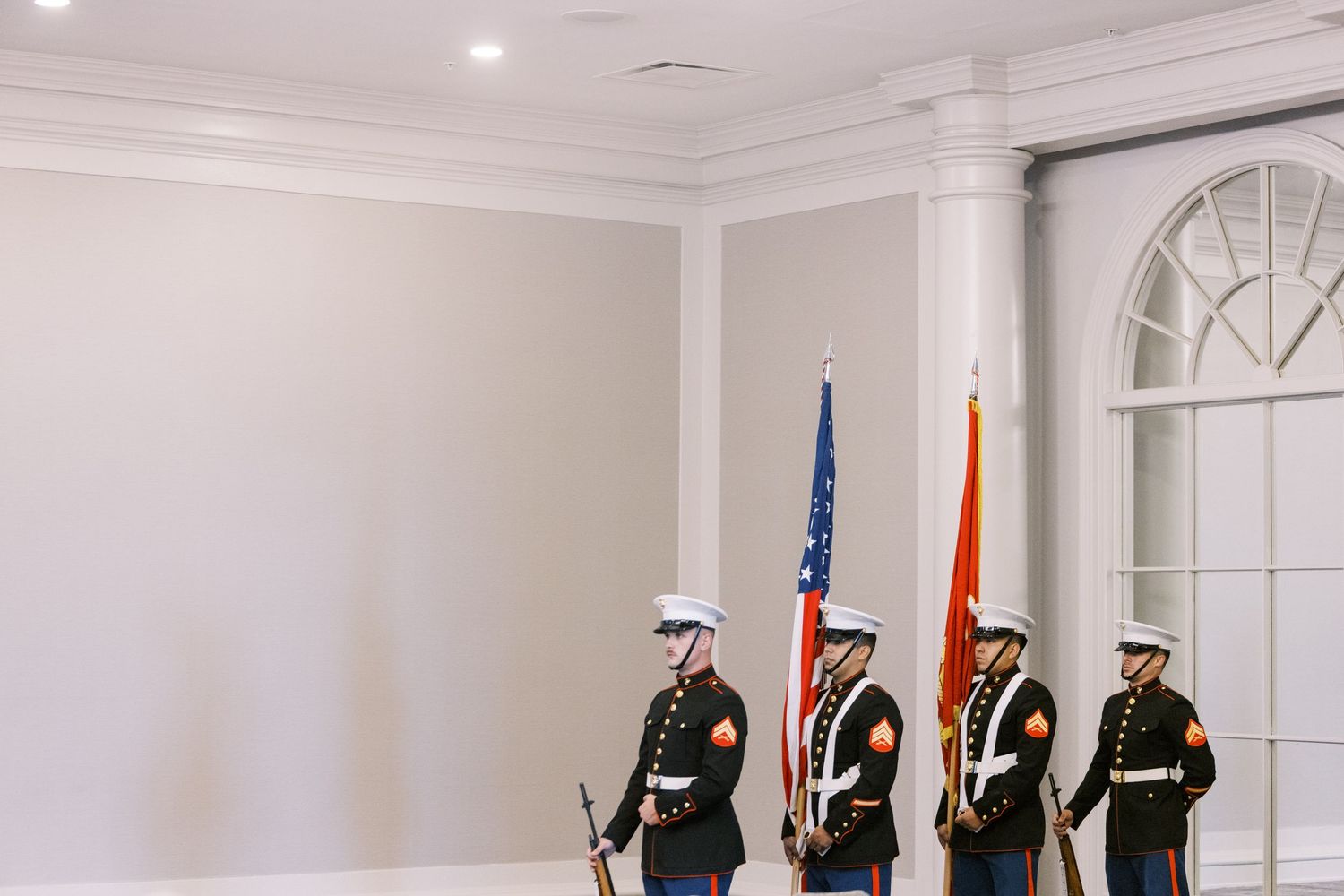 Military honor guard members stand at attention with flags in a formal white room.
