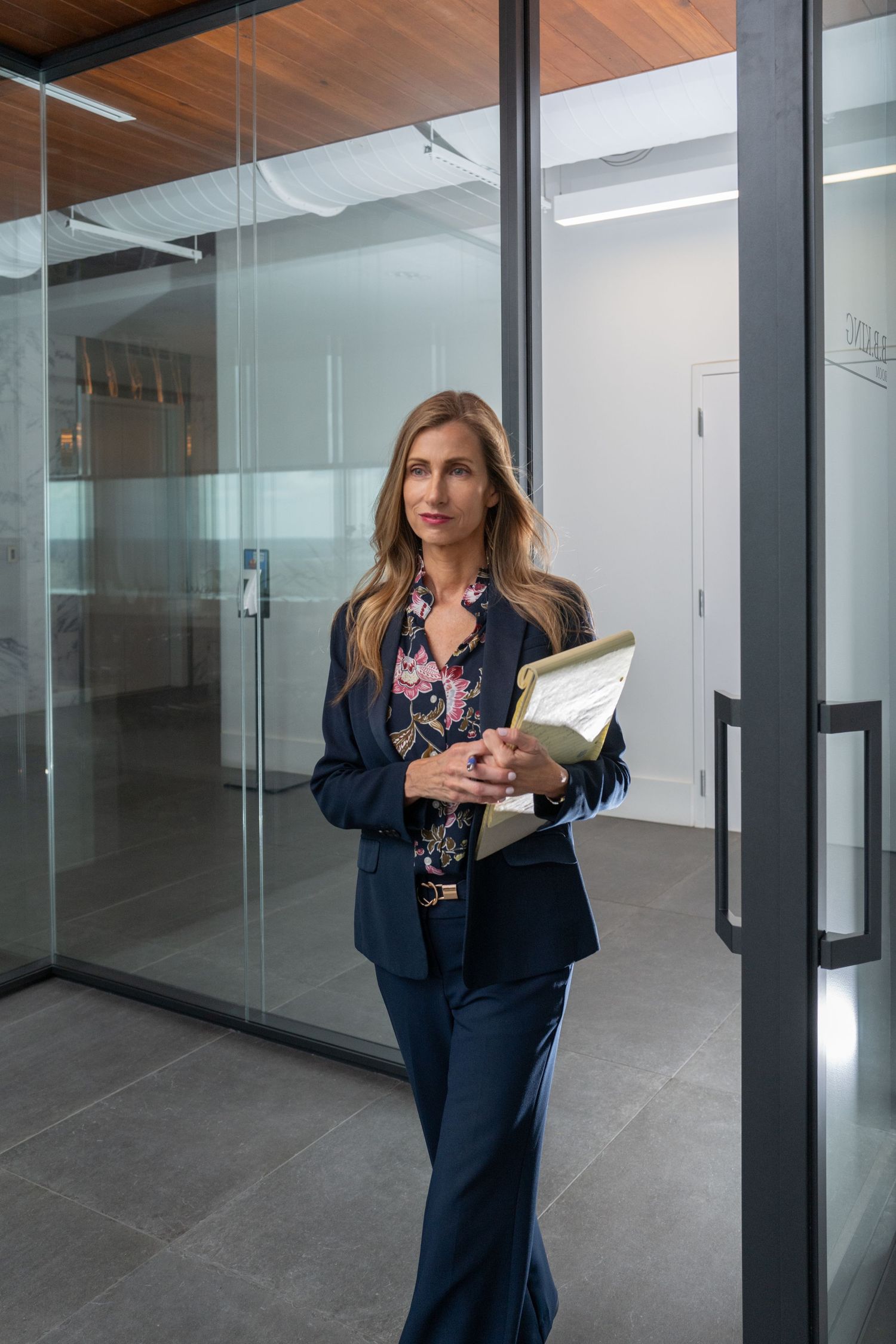 Professional woman in navy suit holding files walks through modern glass office corridor.