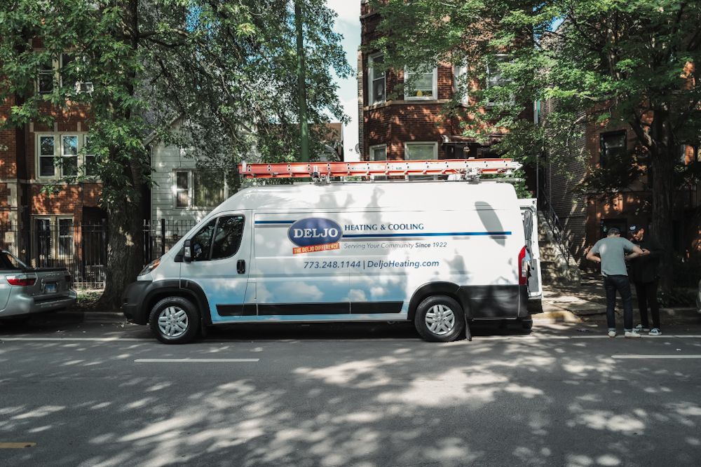 A white HVAC service van parked on a tree-lined urban street with a technician standing nearby.