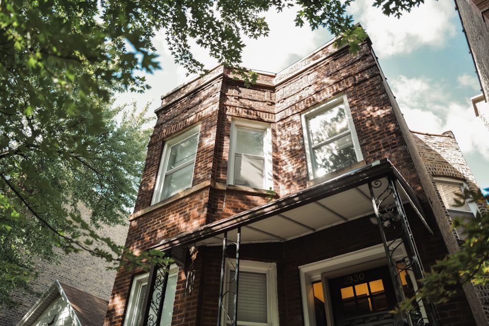 Brick two-story home with bay windows, covered porch, and lush green trees in an urban neighborhood setting.