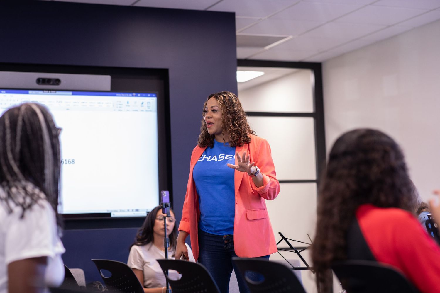 A presenter wearing a pink blazer and blue shirt speaks during a business meeting with a presentation screen visible.