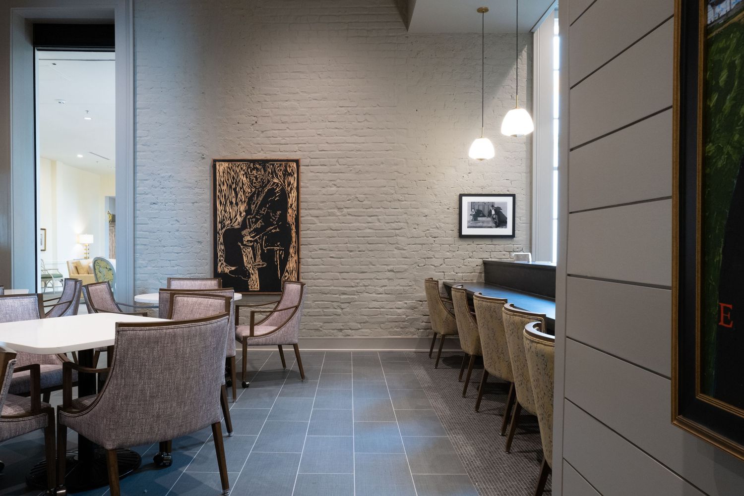 Modern restaurant dining area with upholstered chairs, pendant lights, and artwork on exposed brick wall.