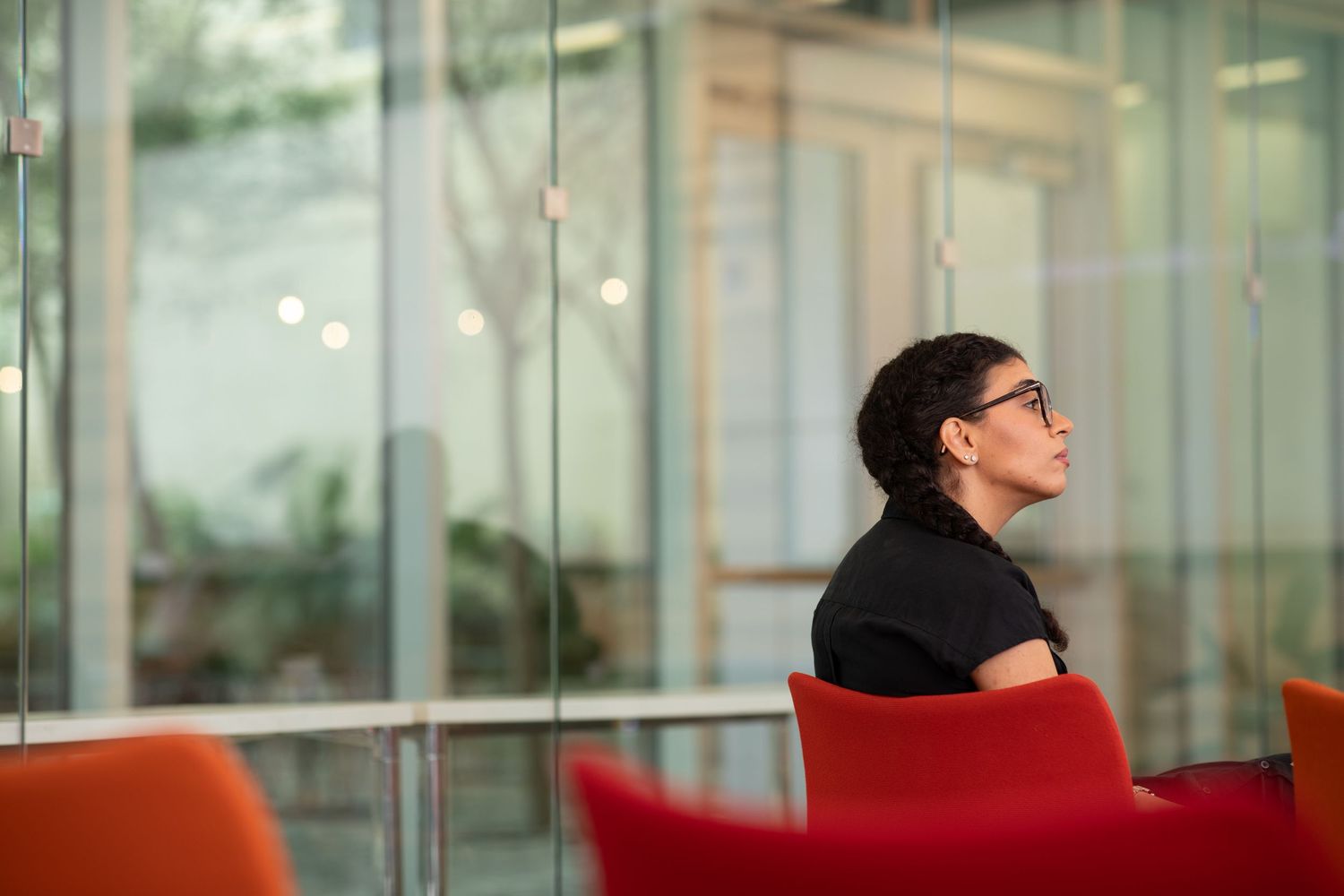 A person in glasses sits on a red couch in a modern office space with glass walls and natural lighting.