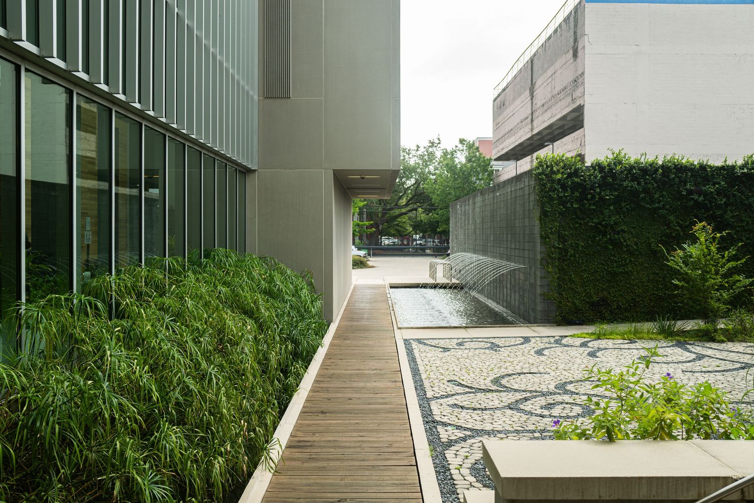 Modern building walkway featuring a wooden deck path alongside green landscaping and geometric stone patterns.