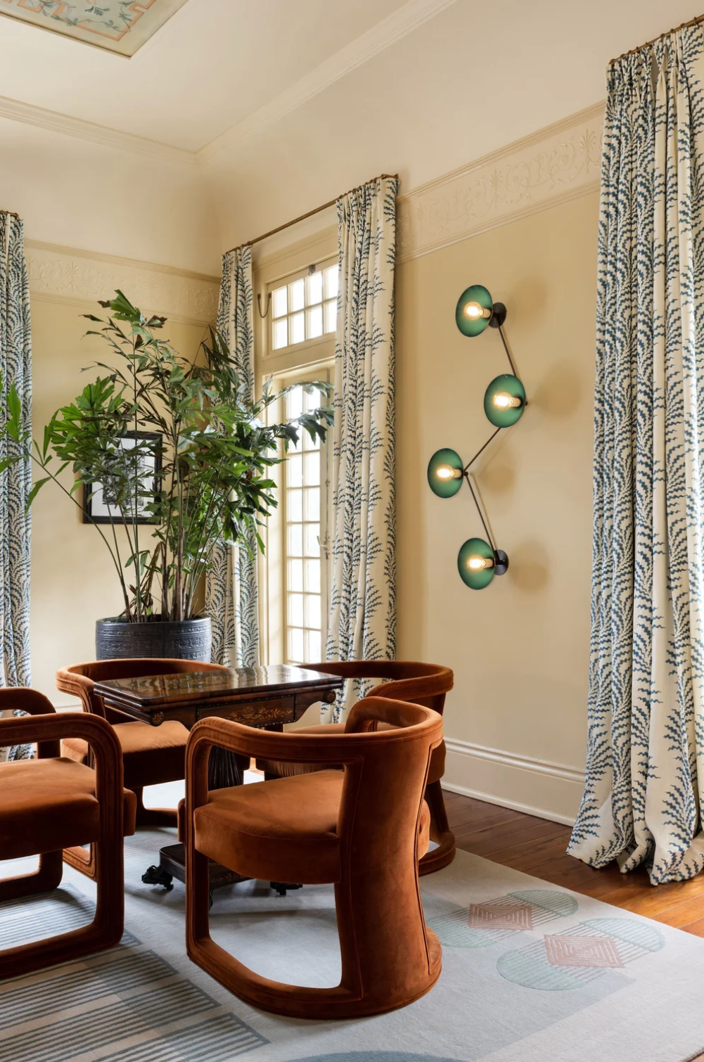 Modern living room interior with brown velvet barrel chairs, patterned curtains, and green wall sconces in natural light.