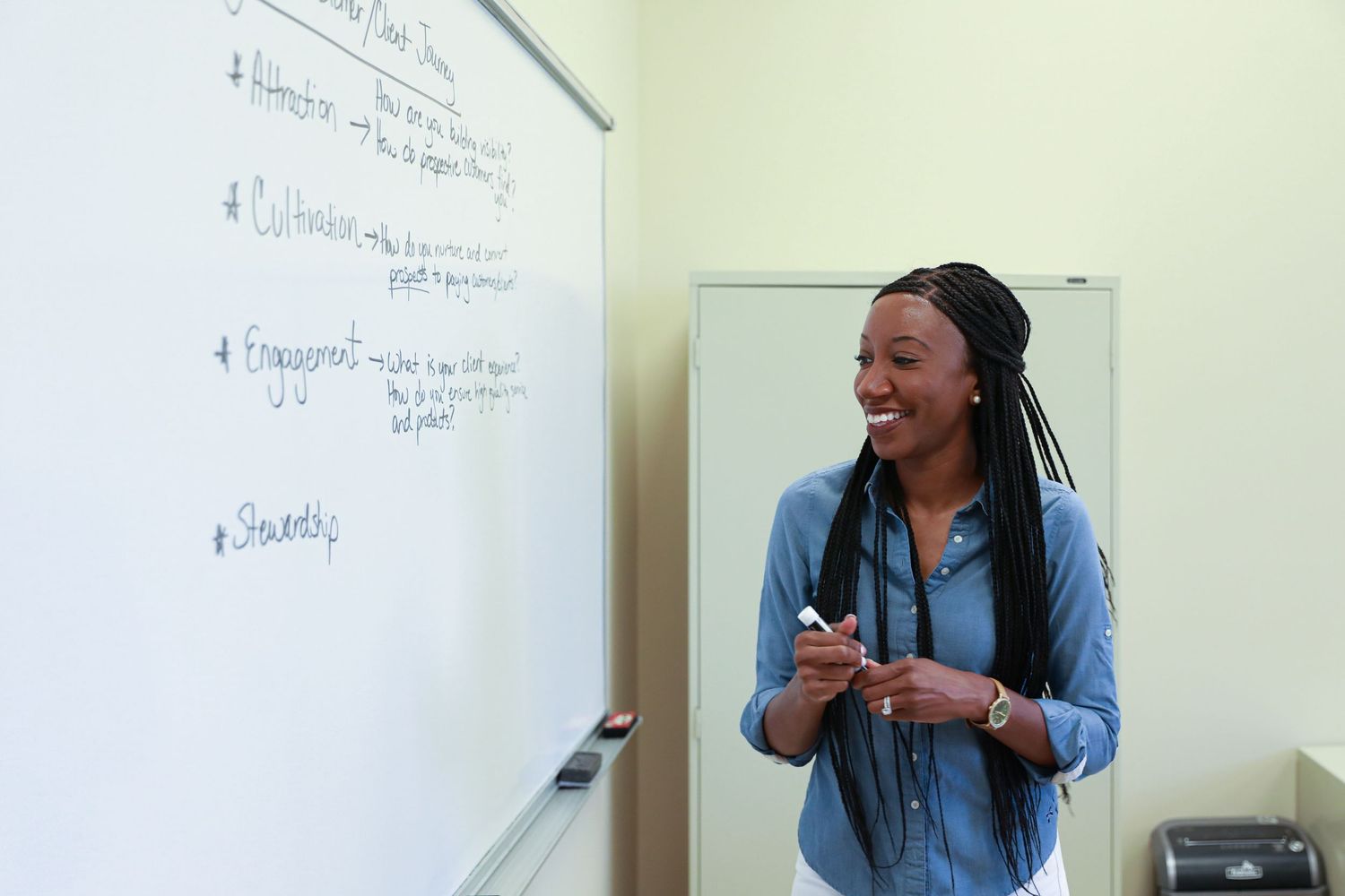 Educator laughs while teaching at whiteboard in classroom setting.