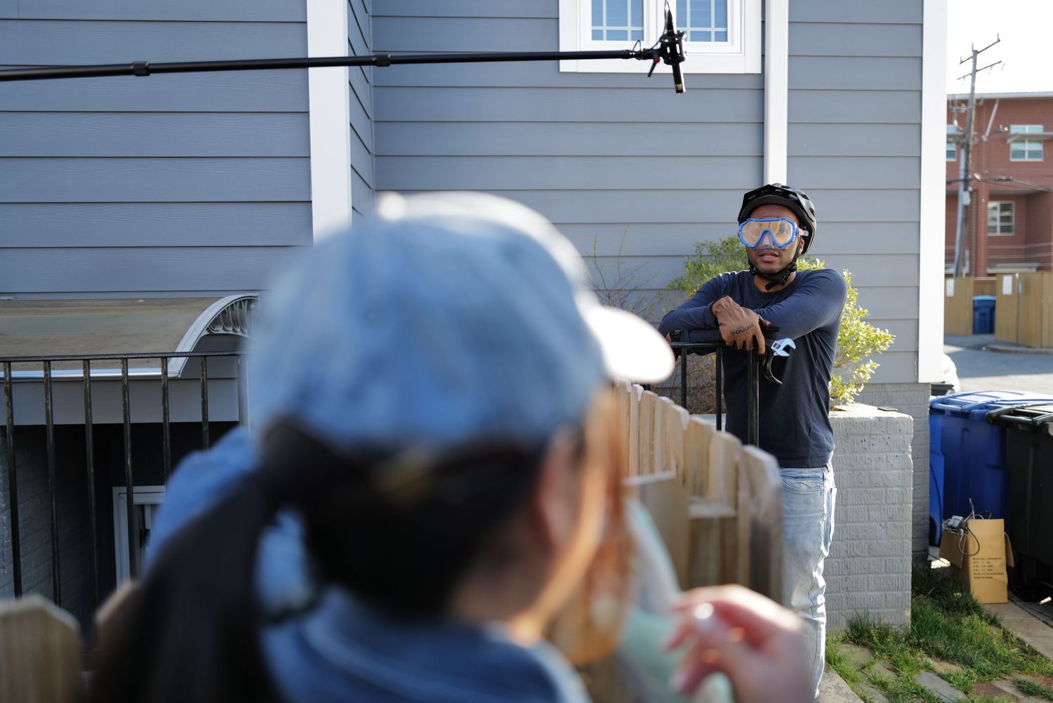 Two people socializing outdoors near a wooden fence beside a gray house on a sunny day.