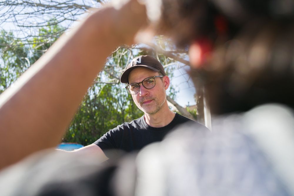Close-up portrait of a man wearing glasses and black cap outdoors in natural sunlight.