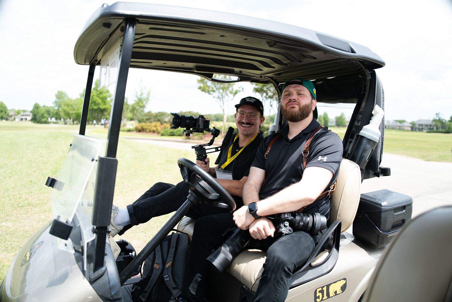 Two men riding in a golf cart on a sunny day at an outdoor event, both wearing black attire.