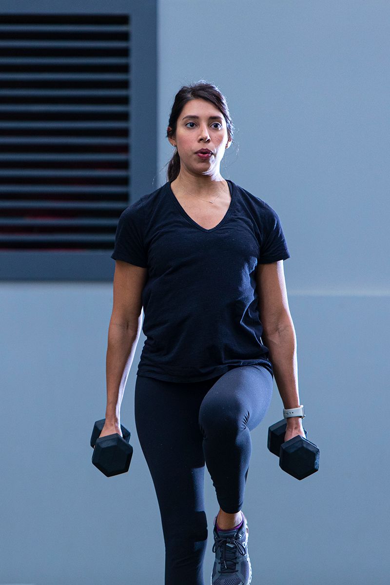 Person in black workout attire performing lunges with dumbbells against blue wall background.