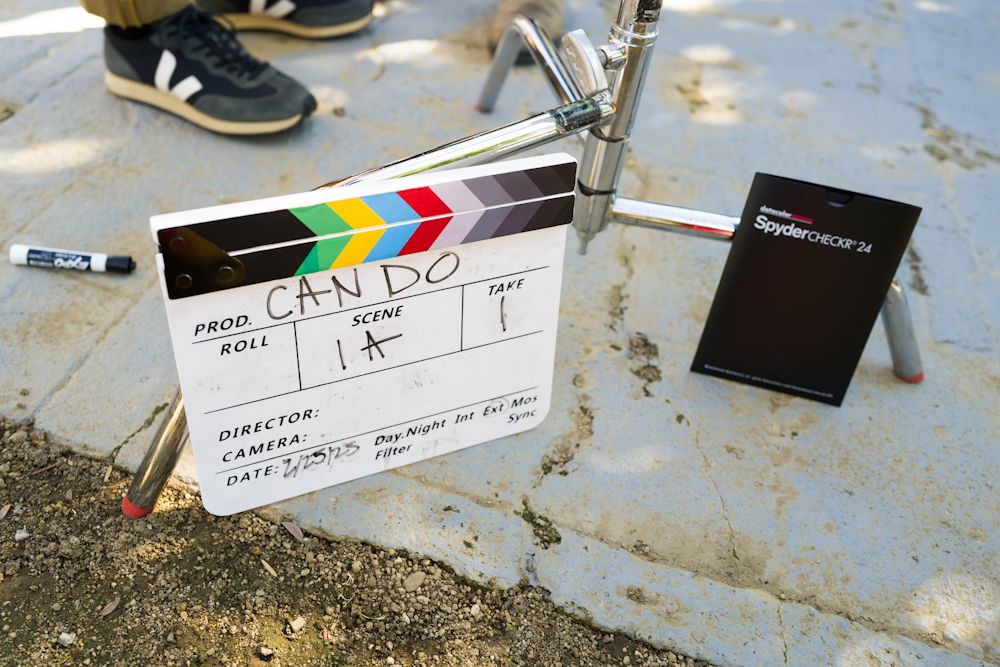 Film clapperboard reading CAN DO on a movie set outdoors, with a SpyderCHECKR color calibration card nearby.
