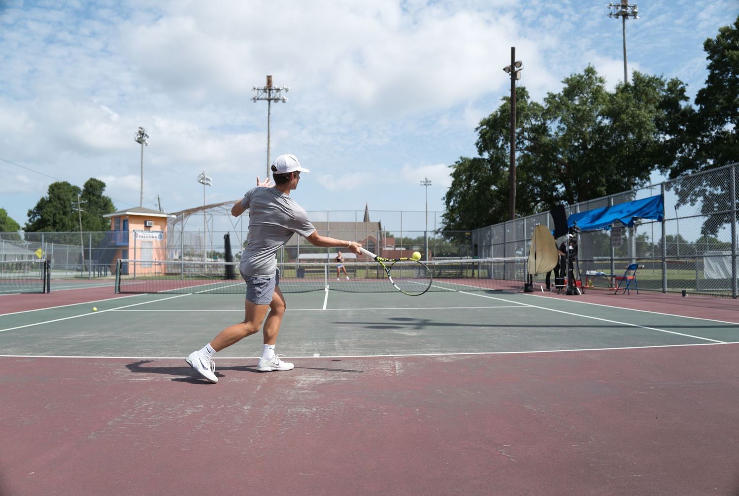 A tennis player mid-swing on an outdoor court on a partly cloudy day.
