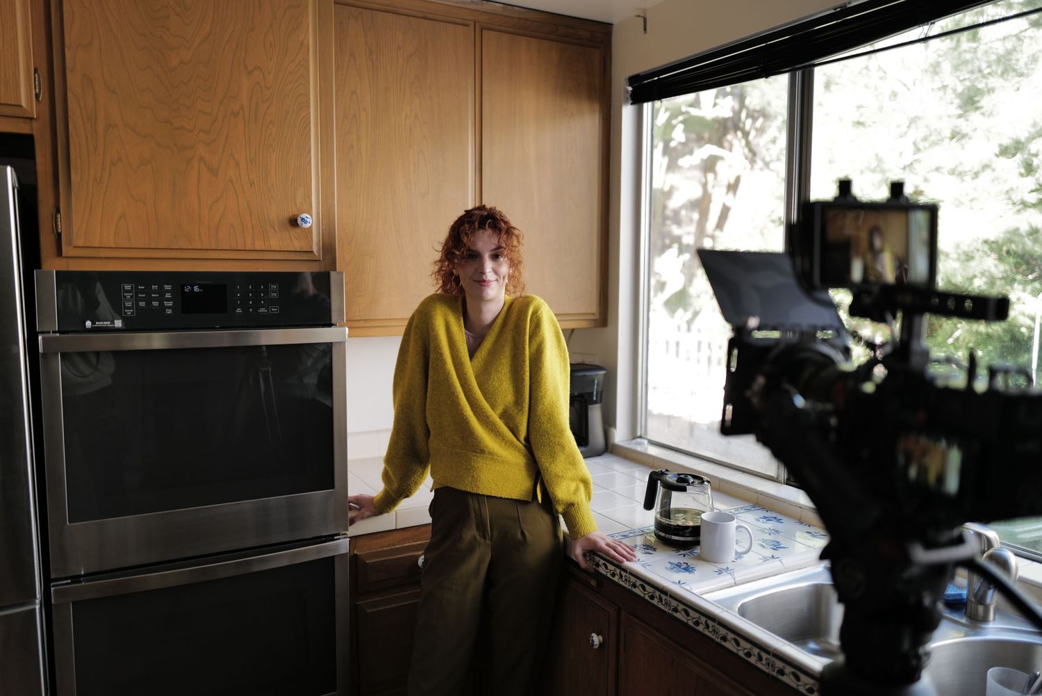 Woman in yellow sweater standing in modern kitchen being filmed by smartphone on tripod near window.